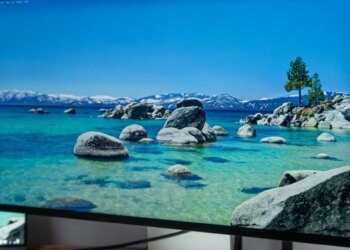 The Apple Studio Display XDR displaying a water landscape on a wooden desk next to a Macbook.