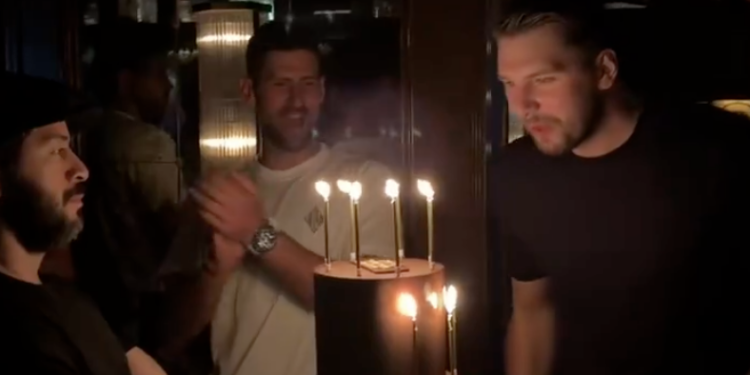 Luka Dončić blowing out birthday candles as Novak Djokovic claps behind him.