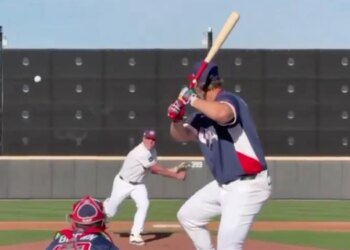 A baseball player in a red, white, and blue uniform batting while a pitcher throws and a catcher crouches.