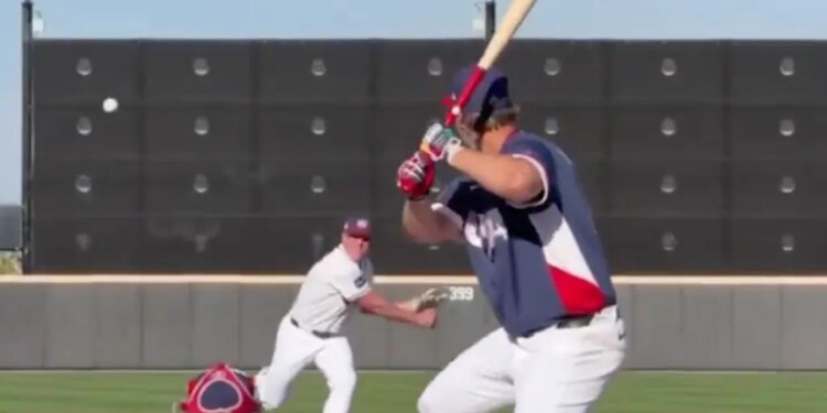 A baseball player in a red, white, and blue uniform batting while a pitcher throws and a catcher crouches.