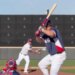 A baseball player in a red, white, and blue uniform batting while a pitcher throws and a catcher crouches.