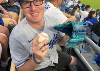 A man in a Dodgers hat and jersey smiling at a baseball game, holding a baseball and a turquoise glove.