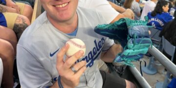 A man in a Dodgers hat and jersey smiling at a baseball game, holding a baseball and a turquoise glove.