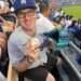 A man in a Dodgers hat and jersey smiling at a baseball game, holding a baseball and a turquoise glove.