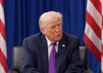 Donald Trump speaking while seated in front of American flags and a blue backdrop.