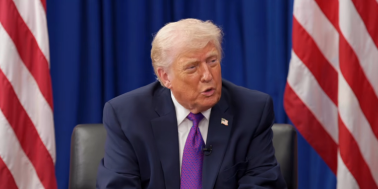 Donald Trump speaking while seated in front of American flags and a blue backdrop.