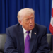 Donald Trump speaking while seated in front of American flags and a blue backdrop.