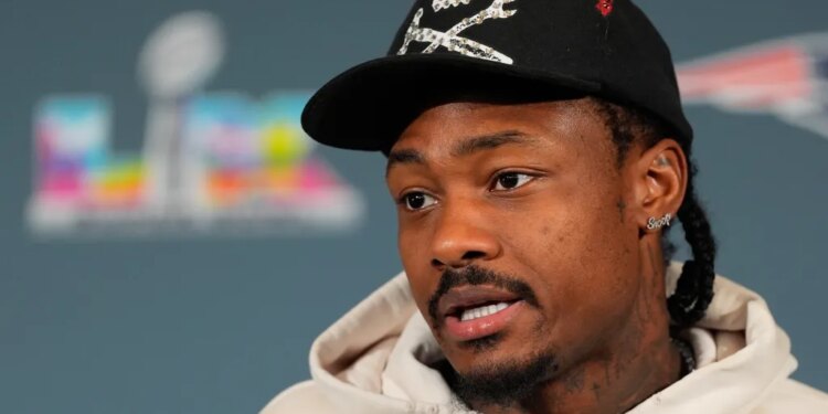 Stefon Diggs speaking, wearing a black cap with a skull and crossbones design, and a light-colored hoodie.