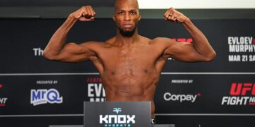 Michael Page posing shirtless on a weigh-in scale, flexing his arms.