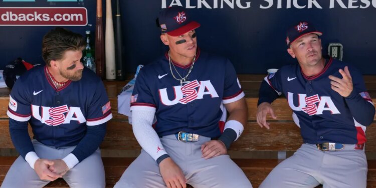 United States' Bryce Harper, left, Aaron Judge, center, and Alex Bregman sit in the team dugout prior to an exhibition baseball game against the Colorado Rockies Wednesday, March 4, 2026, in Scottsdale, Ariz.