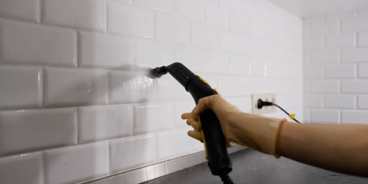 Person using a steam cleaner to clean white subway tiles and grout in a kitchen.