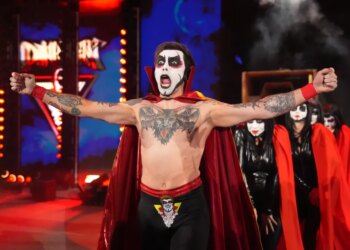 Wrestler Danhausen with painted face and red cape, arms outstretched, with other costumed people behind him.