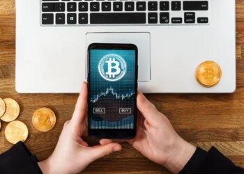Person using a smartphone app to buy or sell Bitcoins, with a laptop and physical Bitcoin coins on a wooden table.