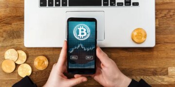 Person using a smartphone app to buy or sell Bitcoins, with a laptop and physical Bitcoin coins on a wooden table.