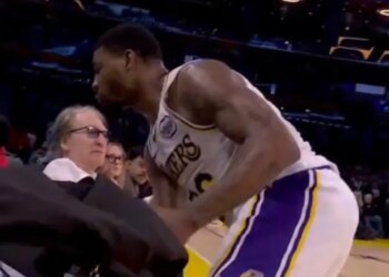 A basketball player in a white and purple uniform bends over near a spectator in glasses.