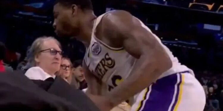 A basketball player in a white and purple uniform bends over near a spectator in glasses.