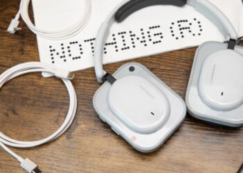 The Nothing Headphone A with its carrying bag, 3.5mm cable, and USB-C cable on a wooden desk with an iPhone nearby.