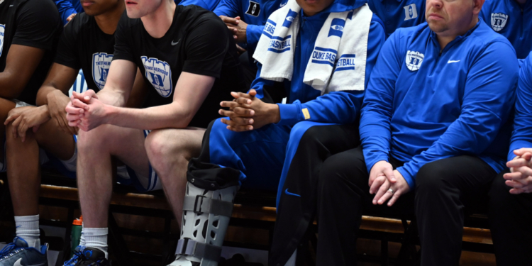 Injured Duke Blue Devils guard Caleb Foster wearing a walking boot on the bench.  Injured Duke Blue Devils guard Caleb Foster (center) looks on with a while wearing a walking boot during the second half against the North Carolina Tar Heels at Cameron Indoor Stadium.
