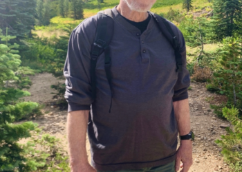 Chuck Norris wearing a black cap, gray shirt, and green shorts, standing on a hiking trail.
