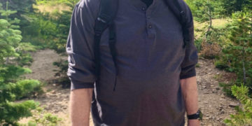 Chuck Norris wearing a black cap, gray shirt, and green shorts, standing on a hiking trail.