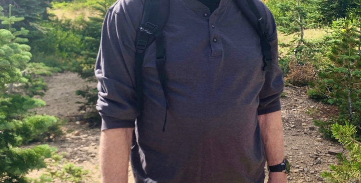 Chuck Norris wearing a black cap, gray shirt, and green shorts, standing on a hiking trail.