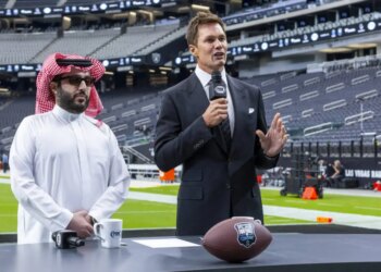 Turki Alalshikh and Tom Brady at a podium with a football in a stadium.