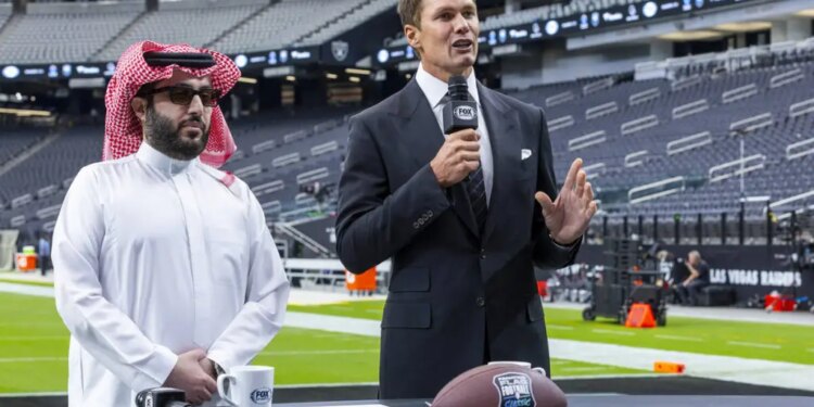 Turki Alalshikh and Tom Brady at a podium with a football in a stadium.