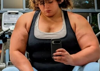 Jammie Booker, a strongwoman competitor, sits in a gym with headphones on, looking down at her phone.