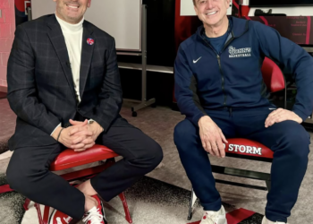 Two men, one in a suit and one in athletic wear, sit on red chairs and smile at the camera.