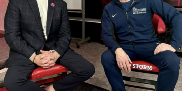 Two men, one in a suit and one in athletic wear, sit on red chairs and smile at the camera.