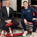 Two men, one in a suit and one in athletic wear, sit on red chairs and smile at the camera.