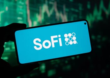 A hand holding a smartphone displaying the SoFi logo on its screen, with a blurry green stock market graph in the background.