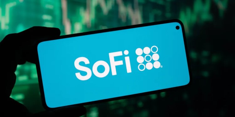 A hand holding a smartphone displaying the SoFi logo on its screen, with a blurry green stock market graph in the background.