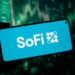 A hand holding a smartphone displaying the SoFi logo on its screen, with a blurry green stock market graph in the background.