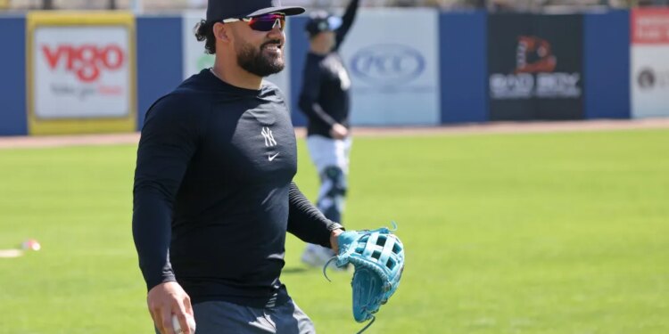 Jasson Domínguez smiling on the field in a Yankees cap and long-sleeve shirt, holding a baseball in one hand and a blue glove in the other.