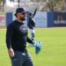 Jasson Domínguez smiling on the field in a Yankees cap and long-sleeve shirt, holding a baseball in one hand and a blue glove in the other.