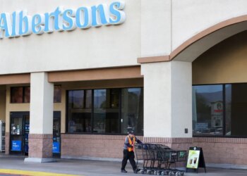 A man in a hi-vis vest pushes a line of shopping carts outside an Albertsons grocery store.