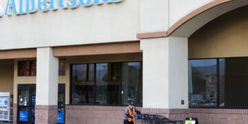 A man in a hi-vis vest pushes a line of shopping carts outside an Albertsons grocery store.