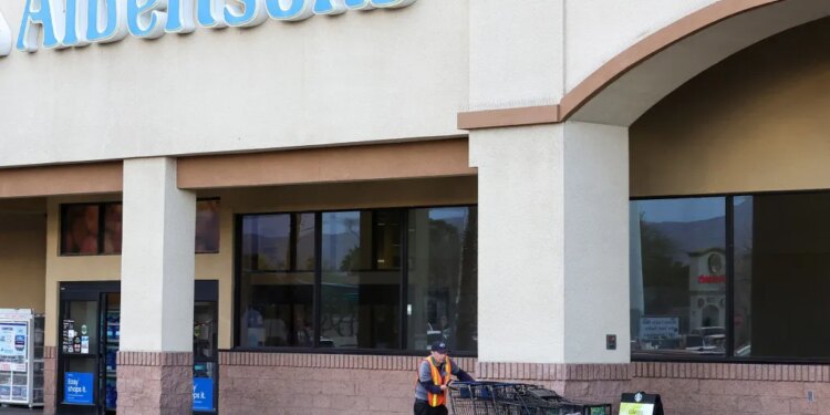 A man in a hi-vis vest pushes a line of shopping carts outside an Albertsons grocery store.
