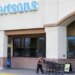A man in a hi-vis vest pushes a line of shopping carts outside an Albertsons grocery store.
