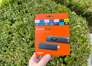 Hand holding an orange Amazon Fire TV Stick streaming device packaging against a green foliage background.
