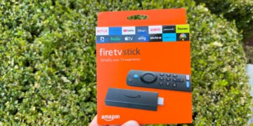Hand holding an orange Amazon Fire TV Stick streaming device packaging against a green foliage background.