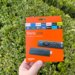 Hand holding an orange Amazon Fire TV Stick streaming device packaging against a green foliage background.