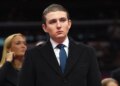 Barron Trump in a dark coat and blue tie, looking forward, with a blurred blonde woman behind him.