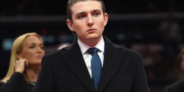 Barron Trump in a dark coat and blue tie, looking forward, with a blurred blonde woman behind him.