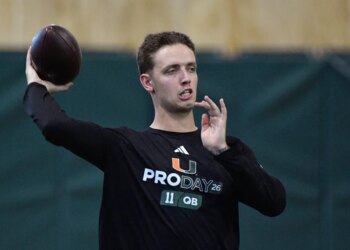 A football player wearing a black "PRO DAY 26 11 QB" long-sleeved shirt, holding a football with his right hand in preparation to throw.