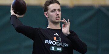 A football player wearing a black "PRO DAY 26 11 QB" long-sleeved shirt, holding a football with his right hand in preparation to throw.