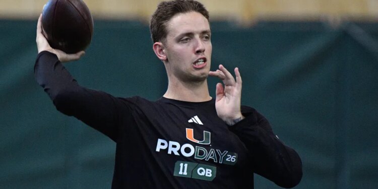 A football player wearing a black "PRO DAY 26 11 QB" long-sleeved shirt, holding a football with his right hand in preparation to throw.