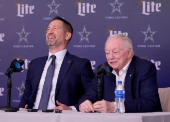 Brian Schottenheimer laughing as Jerry Jones responds to a question at a press conference.