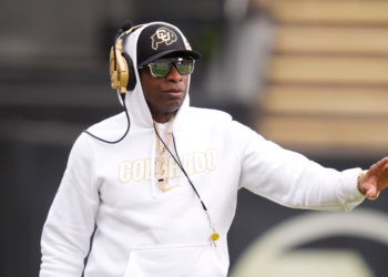 Deion Sanders, head coach of the Colorado Buffaloes, in a white hoodie, hat, sunglasses, and gold headphones, on the field.
